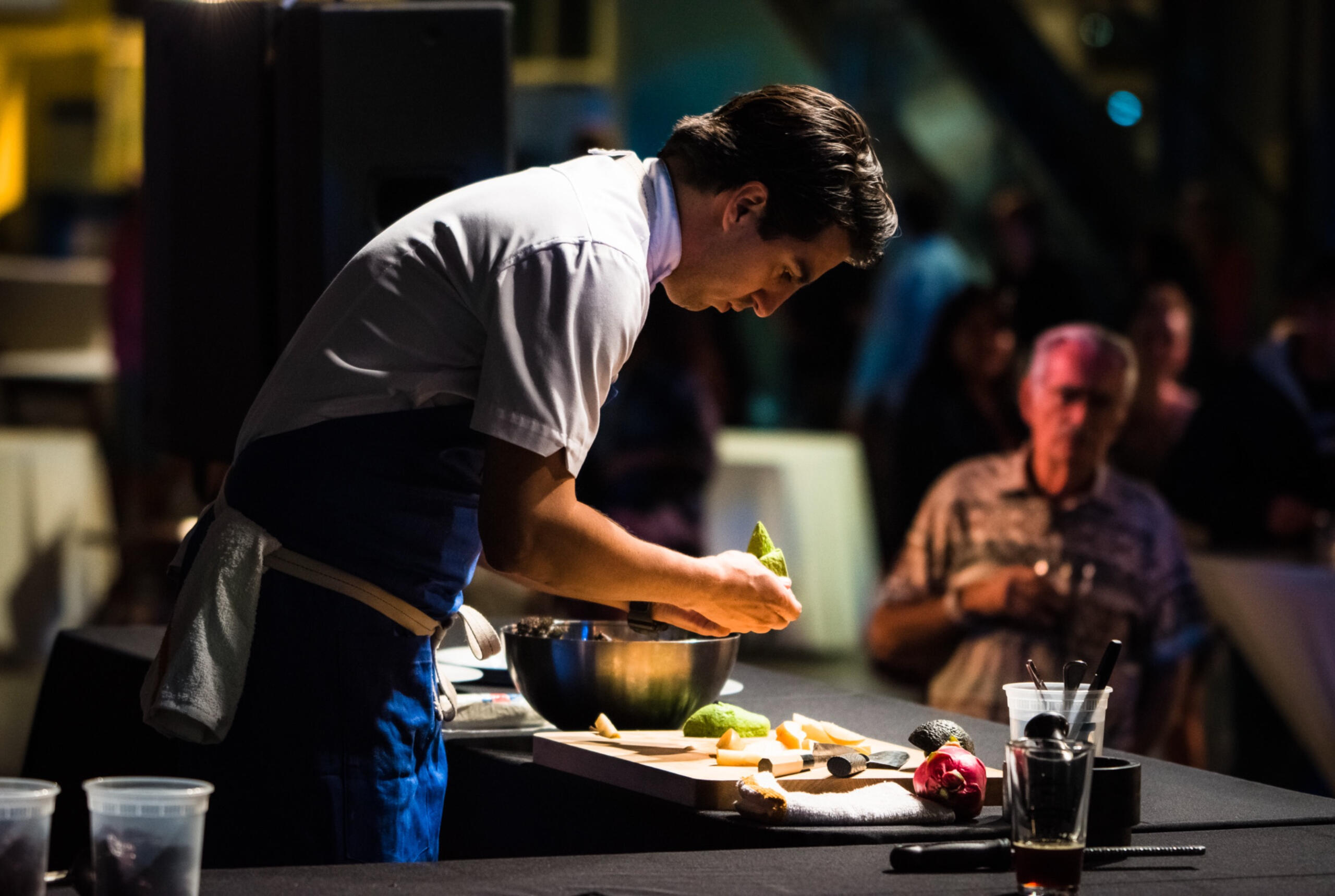 Chef in crisp uniform plating food during a live demo at upscale event, photographed in cinematic lighting with audience engaged in background.